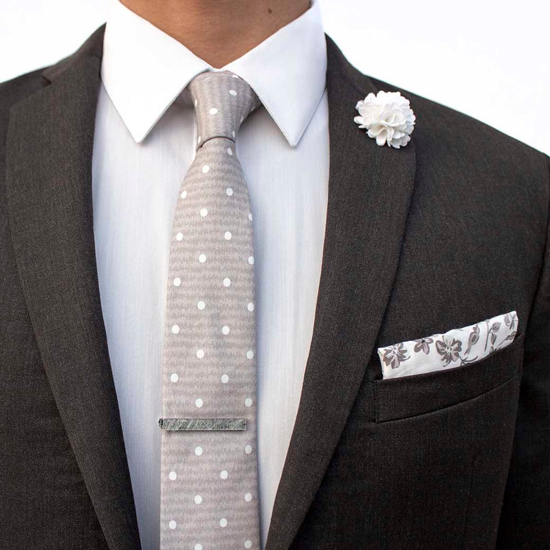 front angle of charcoal suit using menstiel's distorted set of men's suit accessories - A solid white lapel flower with a light floral design on the cotton pocket square is just a start. The grey cotton tie incorporates white polka dots over gray reverbs, and is held together by a fractured glass tie bar.