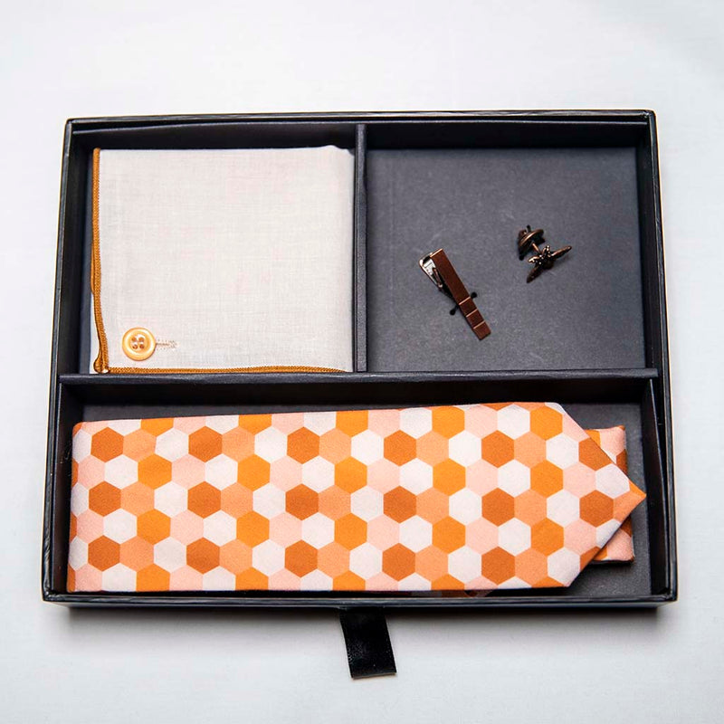 top view of honeycomb set of men's suit accessories in a gift box - A copper bee lapel pin along with a burnished tie bar highlight the multiple shades of rust on the hexagonal cotton tie. We added the double sided linen pocket square with an accentuated border and a sweet button as a finishing touch to the buzz