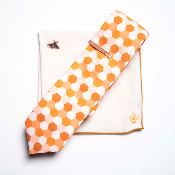 top view of honeycomb set of men's suit accessories - A copper bee lapel pin along with a burnished tie bar highlight the multiple shades of rust on the hexagonal cotton tie. We added the double sided linen pocket square with an accentuated border and a sweet button as a finishing touch to the buzz