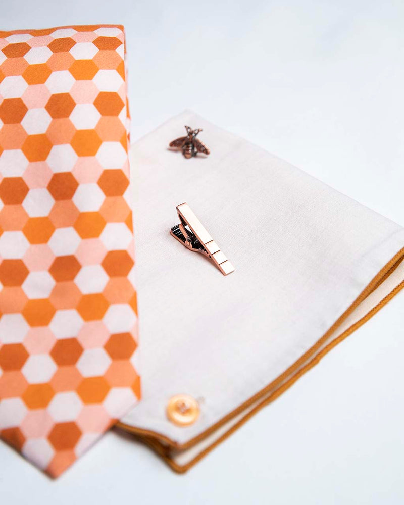 front view of honeycomb set of men's suit accessories - A copper bee lapel pin along with a burnished tie bar highlight the multiple shades of rust on the hexagonal cotton tie. We added the double sided linen pocket square with an accentuated border and a sweet button as a finishing touch to the buzz